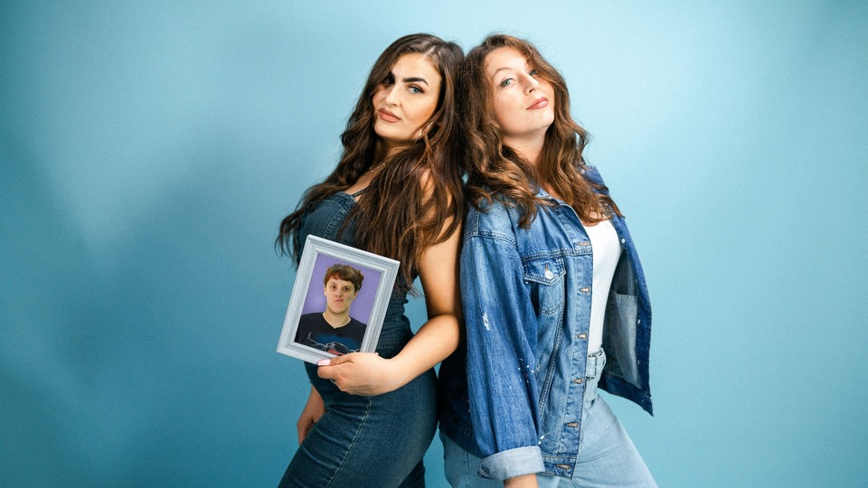 Karina und Larissa stehen mit dem Rücken zueinander vor einem blauen Hintergrund und halten ein Bild von Kaddi in der Hand.